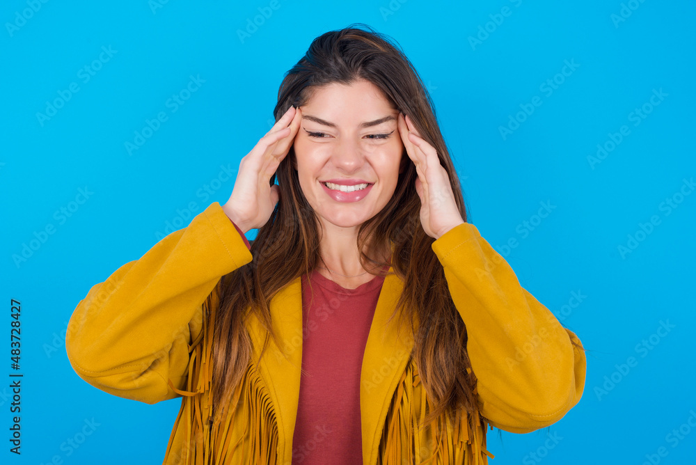Fototapeta premium young brunette woman wearing yellow fringed jacket over blue background concentrating hard on an idea with a serious look, thinking with both index fingers pointing to forehead.