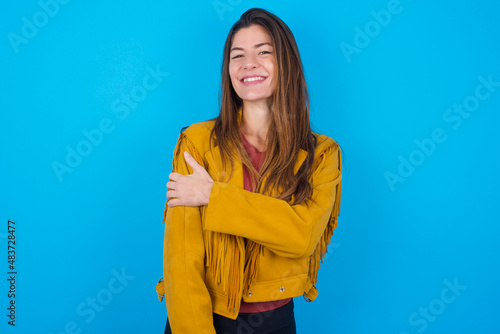 People, lifestyle, youth and happiness concept. Shy pretty young caucasian woman wearing yellow fringed jacket over blue background, feeling happy hugging herself.