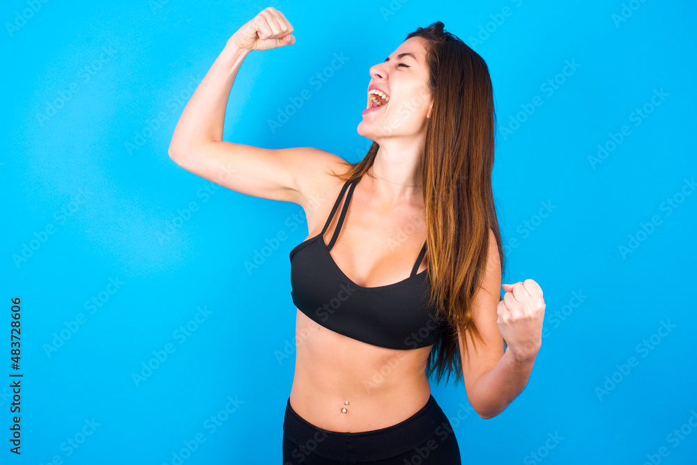 Attractive Young beautiful sportswoman doing sport wearing sportswear over blue background celebrating a victory punching the air with his fists and a beaming toothy smile Attractive Young beautiful sportswoman doing sport wearing sportswear over blue background celebrating a victory punching the air with his fists and a beaming toothy smile