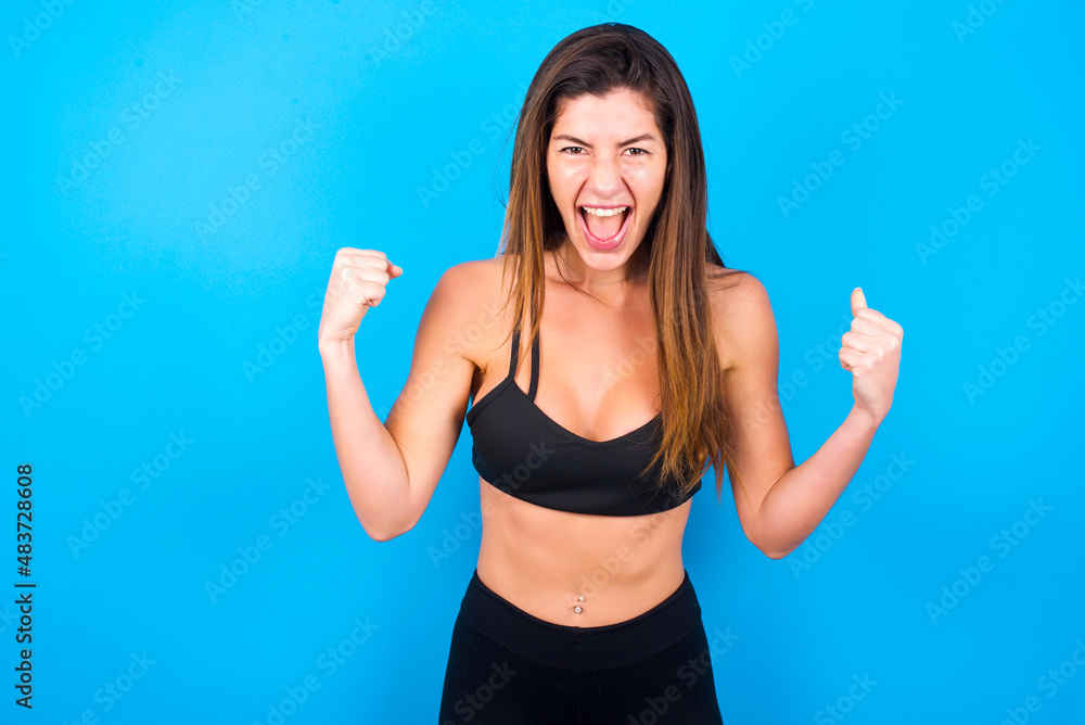 Portrait of Young beautiful sportswoman doing sport wearing sportswear over blue looks with excitement at camera, keeps hands raised over head, notices something unexpected reacts on sudden news. Portrait of Young beautiful sportswoman doing sport wearing sportswear over blue looks with excitement at camera, keeps hands raised over head, notices something unexpected reacts on sudden news.