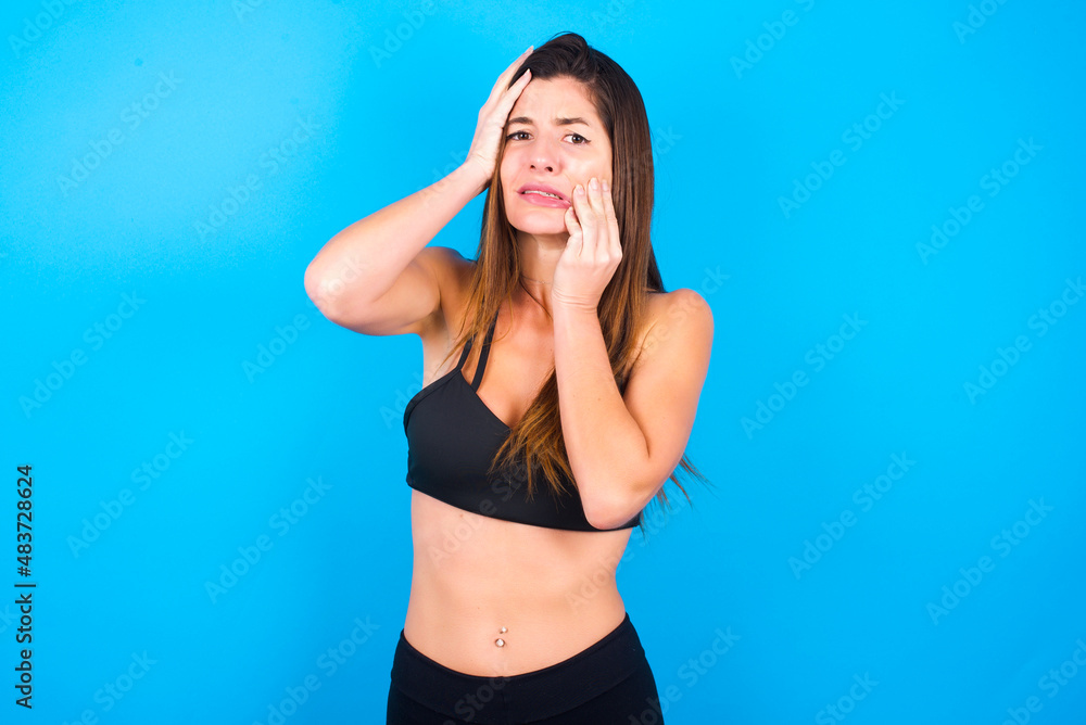 Fototapeta premium Young gloomy Young beautiful sportswoman doing sport wearing sportswear over blue background, hiding face with hands pouting and crying, standing upset and depressed complaining about job problem.