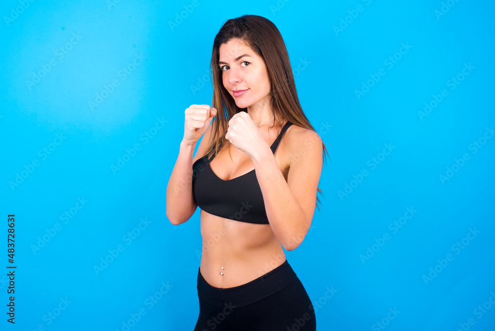 Portrait of attractive Young beautiful sportswoman doing sport wearing sportswear over blue background holding hands in front of him in boxing position going to fight. Portrait of attractive Young beautiful sportswoman doing sport wearing sportswear over blue background holding hands in front of him in boxing position going to fight.