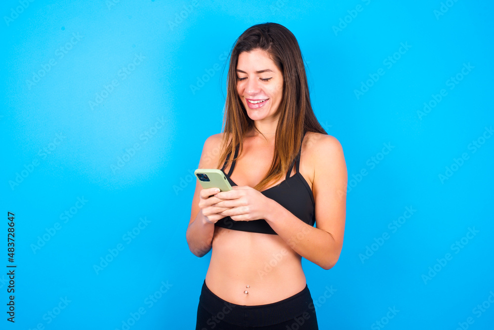 Smiling Young beautiful sportswoman doing sport wearing sportswear using cell phone, messaging, being happy to text with friends, looking at smartphone. Modern technologies and communication. Smiling Young beautiful sportswoman doing sport wearing sportswear using cell phone, messaging, being happy to text with friends, looking at smartphone. Modern technologies and communication.