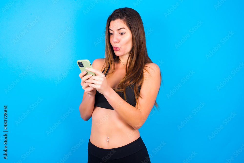Young beautiful sportswoman doing sport wearing sportswear over blue background looks with bugged eyes, holds modern smart phone, receives unexpected message from friend, reads reminder. Young beautiful sportswoman doing sport wearing sportswear over blue background looks with bugged eyes, holds modern smart phone, receives unexpected message from friend, reads reminder.