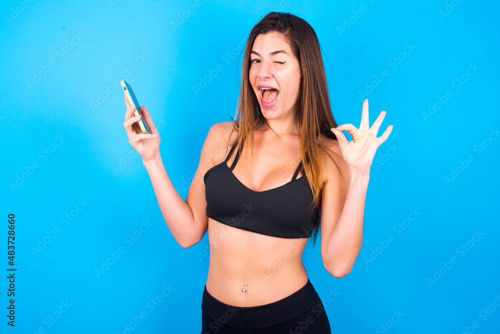 Happy Young beautiful sportswoman doing sport wearing sportswear over blue background sending a message on his smartphone or taking a selfie and making ok sign with his hand. Happy Young beautiful sportswoman doing sport wearing sportswear over blue background sending a message on his smartphone or taking a selfie and making ok sign with his hand.