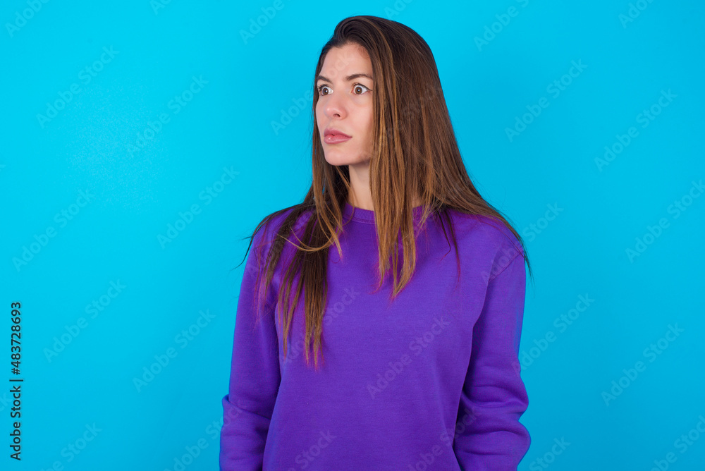 Fototapeta premium young beautiful caucasian woman wearing purple sweater against blue background stares aside with wondered expression has speechless expression. Embarrassed model looks in surprise