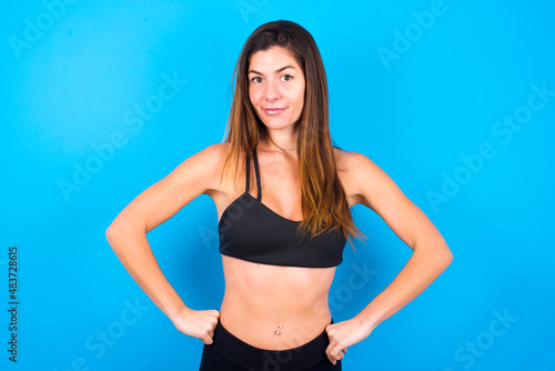 Funny frustrated Young beautiful sportswoman doing sport wearing sportswear over blue background holding hands on waist and silly looking at awkward situation.