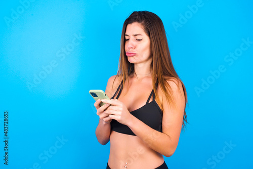 Portrait of a confused Young beautiful sportswoman doing sport wearing sportswear over blue background holding mobile phone and shrugging shoulders and frowning face.