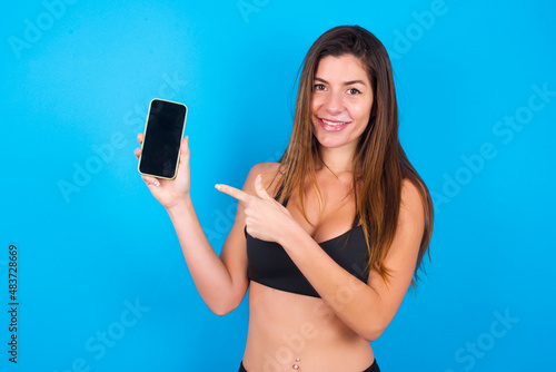 Smiling Young beautiful sportswoman doing sport wearing sportswear over blue background showing and pointing at empty phone screen. Advertisement and communication concept.