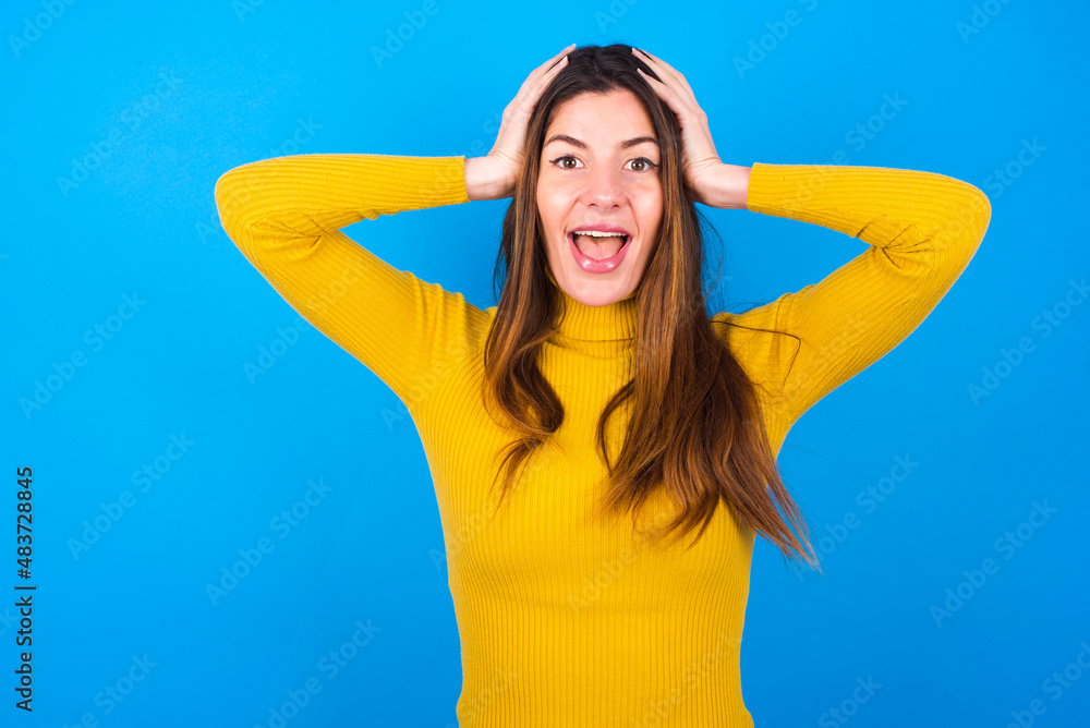 Cheerful overjoyed young woman wearing yellow turtleneck sweater against blue background reacts rising hands over head after receiving great news. Cheerful overjoyed young woman wearing yellow turtleneck sweater against blue background reacts rising hands over head after receiving great news.