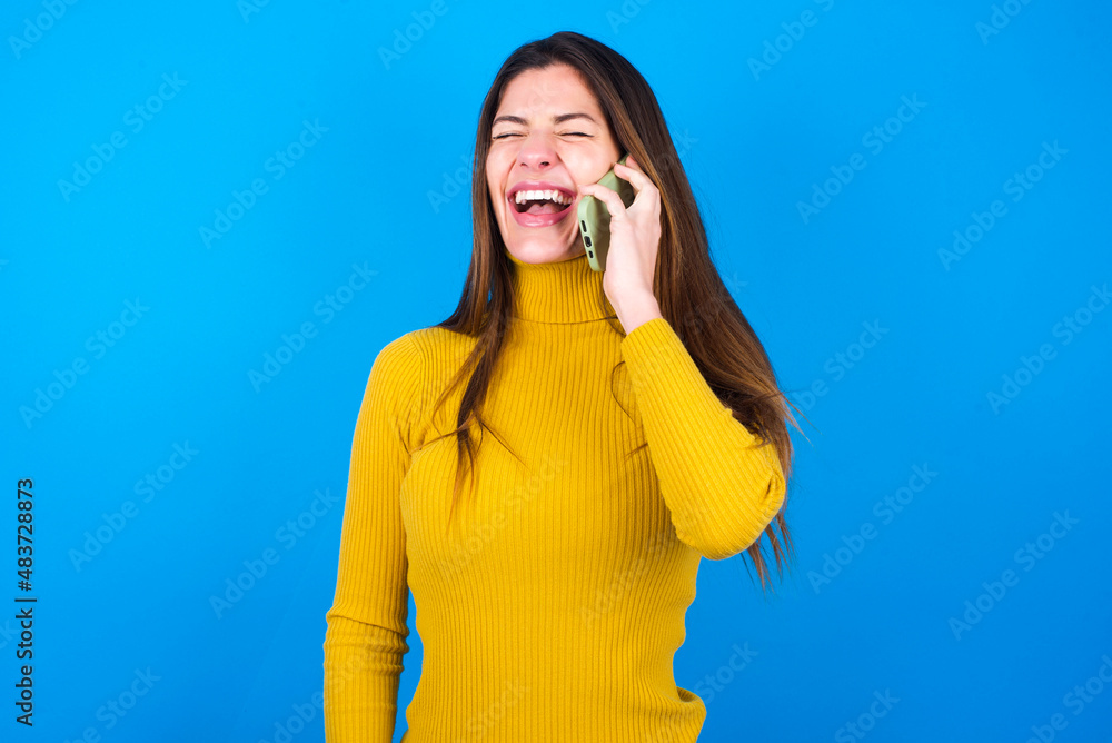 Overemotive happy young woman wearing yellow turtleneck sweater against blue background laughs out positively hears funny story from friend during telephone conversation Overemotive happy young woman wearing yellow turtleneck sweater against blue background laughs out positively hears funny story from friend during telephone conversation