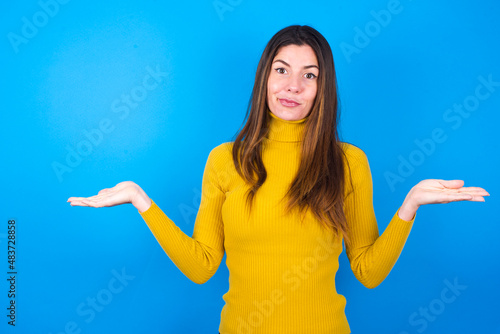 Принти на полотні Careless attractive young woman wearing yellow turtleneck sweater against blue background shrugging shoulders, oops