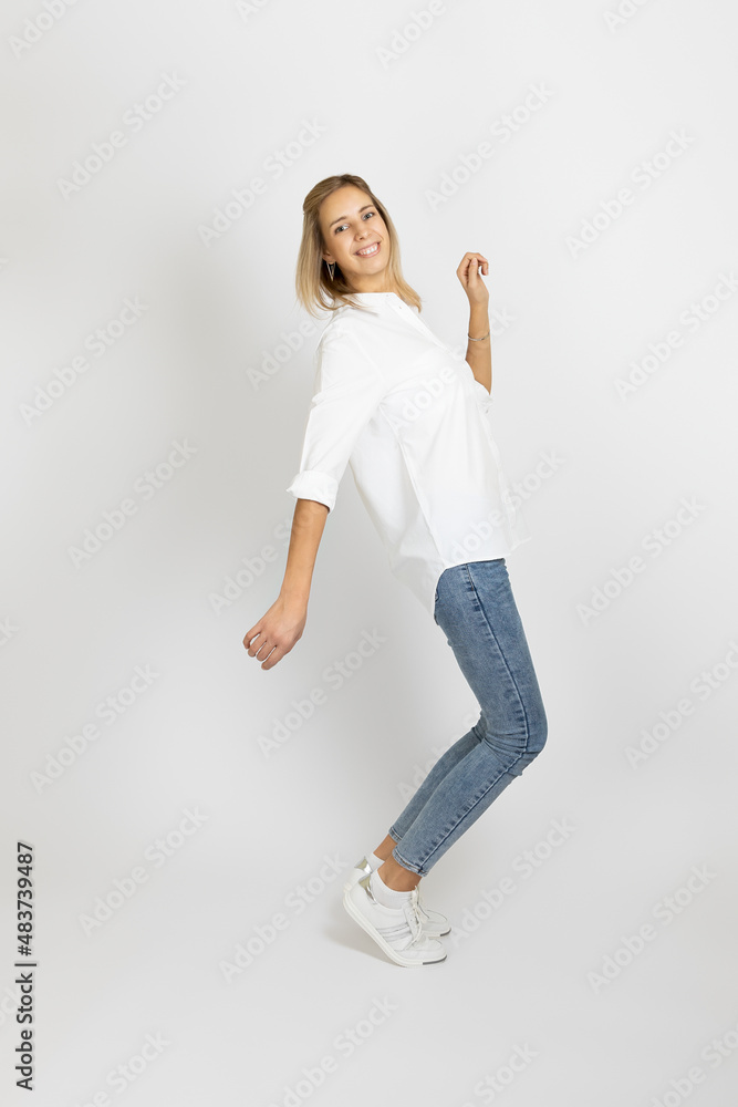 Shining young woman with smile on face posing on white studio background looking at camera wearing white blouse, blue jeans and sneakers. Showing positive emotions. Shining young woman with smile on face posing on white studio background looking at camera wearing white blouse, blue jeans and sneakers. Showing positive emotions.