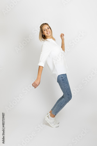 Shining young woman with smile on face posing on white studio background looking at camera wearing white blouse, blue jeans and sneakers. Showing positive emotions. 