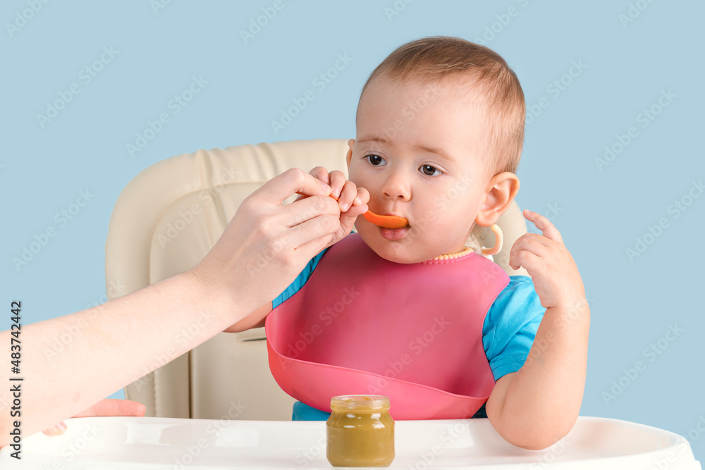 Mom feeds a newborn baby 12-17 months with a spoon mashed broccoli from a glass jar Mom feeds a newborn baby 12-17 months with a spoon mashed broccoli from a glass jar