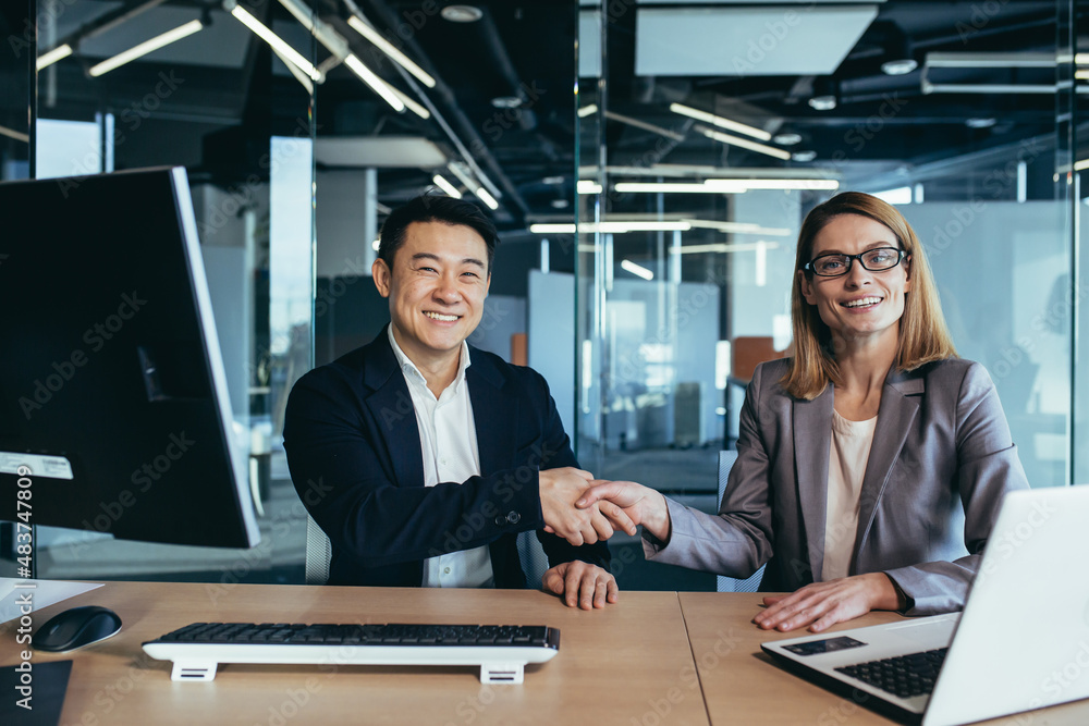 Two workers shaking hands, looking at the camera and smiling, Asian man and woman business people working in a modern office Two workers shaking hands, looking at the camera and smiling, Asian man and woman business people working in a modern office