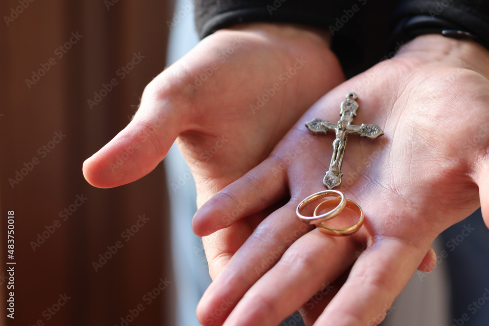 Two gold wedding rings held in the palm of a young man's hand next to a crucifix, lit with natural light