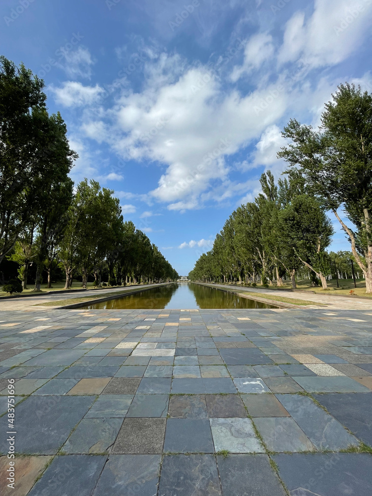 A paved alley in the park with a square pond in the middle surrounded by trees under the blue sky with puffy clouds in summer