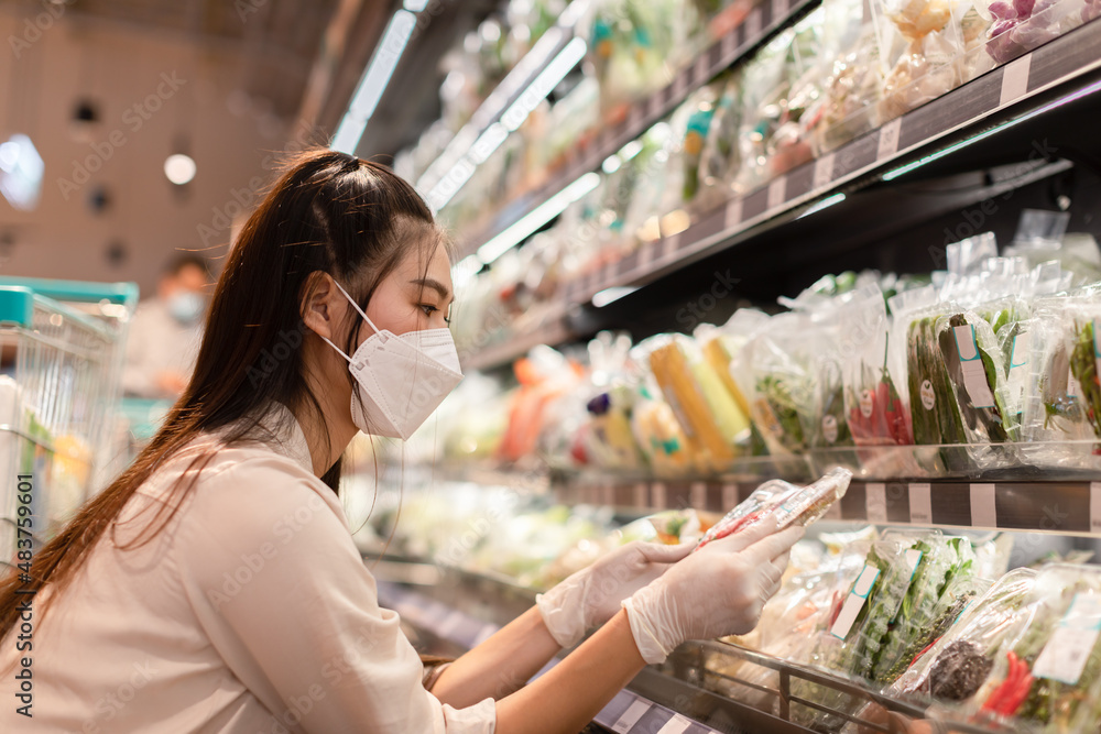 Beautiful asian woman wearing medical face mask and rubber gloves choosing vegetable and fruits on shelf in supermarket.Female looking and hold vegetable shopping in grocery department store