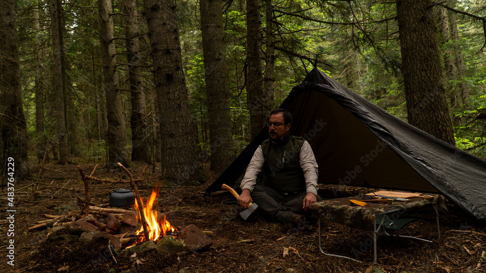 camper man sitting under tarp with ax in dark forest and watching fire man camping  bushcraft in dark pine forest Stock Photo
