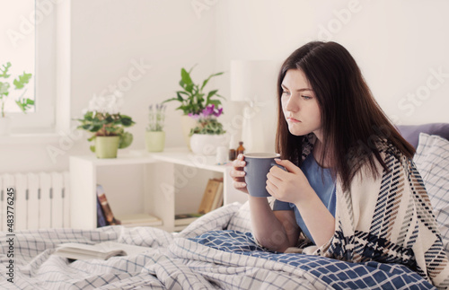 sick teen girl with cup of tea on bed in white room