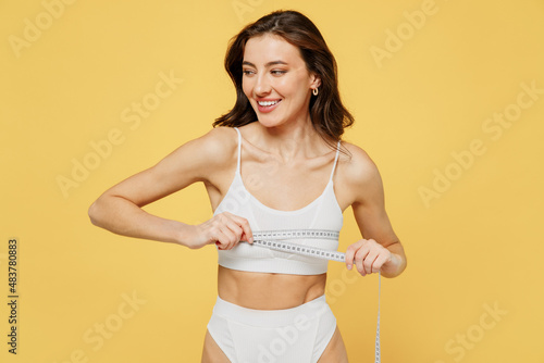 Lovely sexy attractive young brunette woman 20s in white underwear with perfect fit body standing posing hold measure tape on breast look aside on copy space area isolated on plain yellow background © ViDi Studio