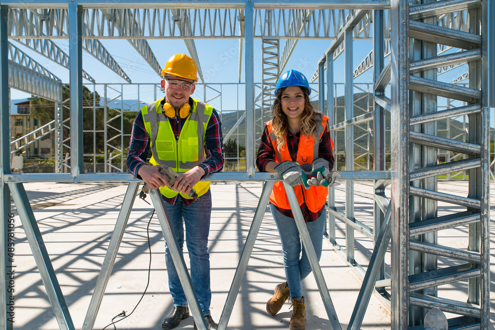 Cheerful construction workers smiling at the camera while sitting. Beautiful young woman and handsome man, sitting in front of framework of the new building.