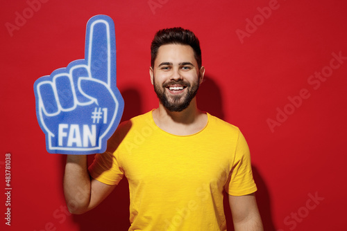 Smiling excited overjoyed young bearded man 20s in yellow t-shirt cheer up support favorite sport team look camera hold fan foam glove finger up isolated on plain dark red background studio portrait. photo