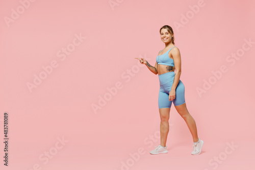 Full size fun young strong sporty fitness trainer woman wear blue tracksuit spend time in home gym walk go point finger aside on area isolated on pastel plain pink background. Workout sport concept.