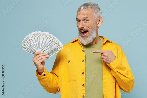 Elderly gray-haired mustache bearded man 50s in yellow shirt hold poitn finger on fan of cash money in dollar banknotes isolated on plain pastel light blue background studio People lifestyle concept photo