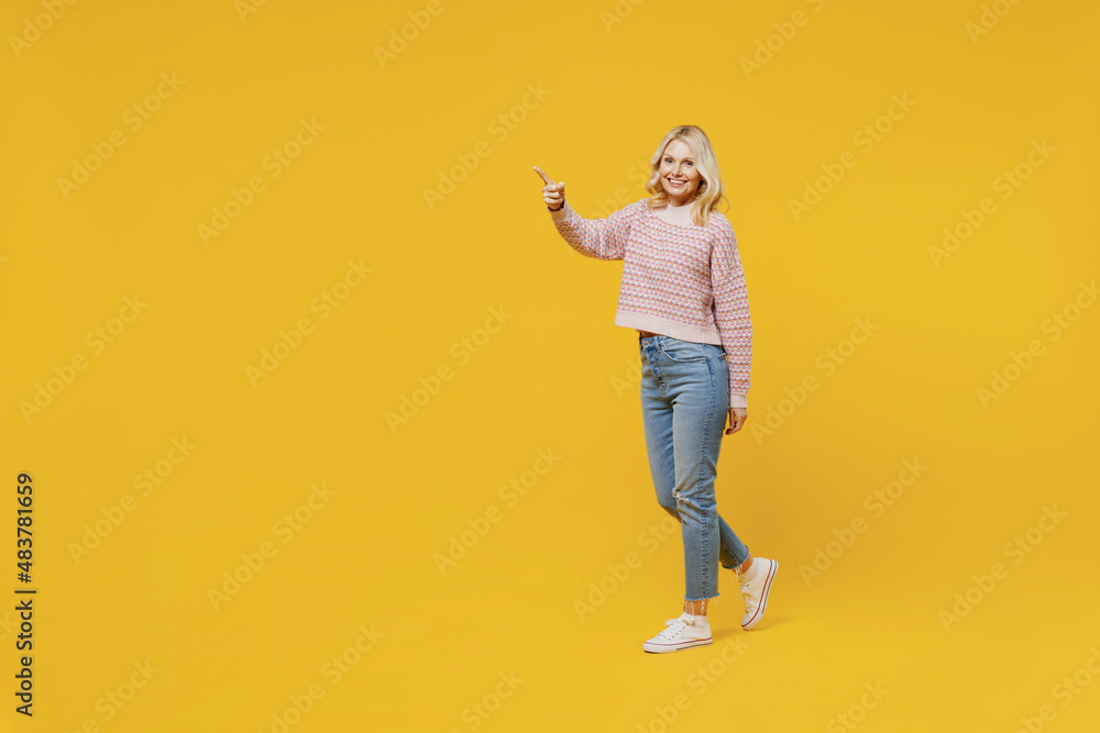 Full body side view happy fun elderly smiling blonde caucasian woman 50s in pink sweater walking going point index finger aside on workspace isolated on plain yellow color background studio portrait. Full body side view happy fun elderly smiling blonde caucasian woman 50s in pink sweater walking going point index finger aside on workspace isolated on plain yellow color background studio portrait.