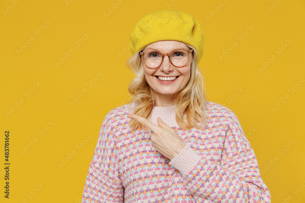 Elderly smiling fashionable positive happy blonde fun woman 50s in pink sweater beret hat glasses point finger aside on workspace isolated on plain yellow background studio. People lifestyle concept