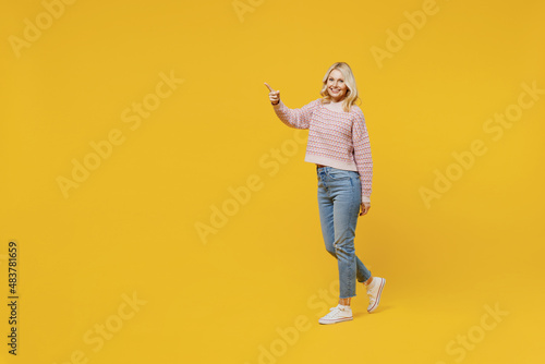 Full body side view happy fun elderly smiling blonde caucasian woman 50s in pink sweater walking going point index finger aside on workspace isolated on plain yellow color background studio portrait.