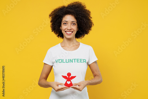 Young woman of African American ethnicity wears white volunteer t-shirt hold in hands little paper angel isolated on plain yellow background. Voluntary free work assistance help charity grace concept.