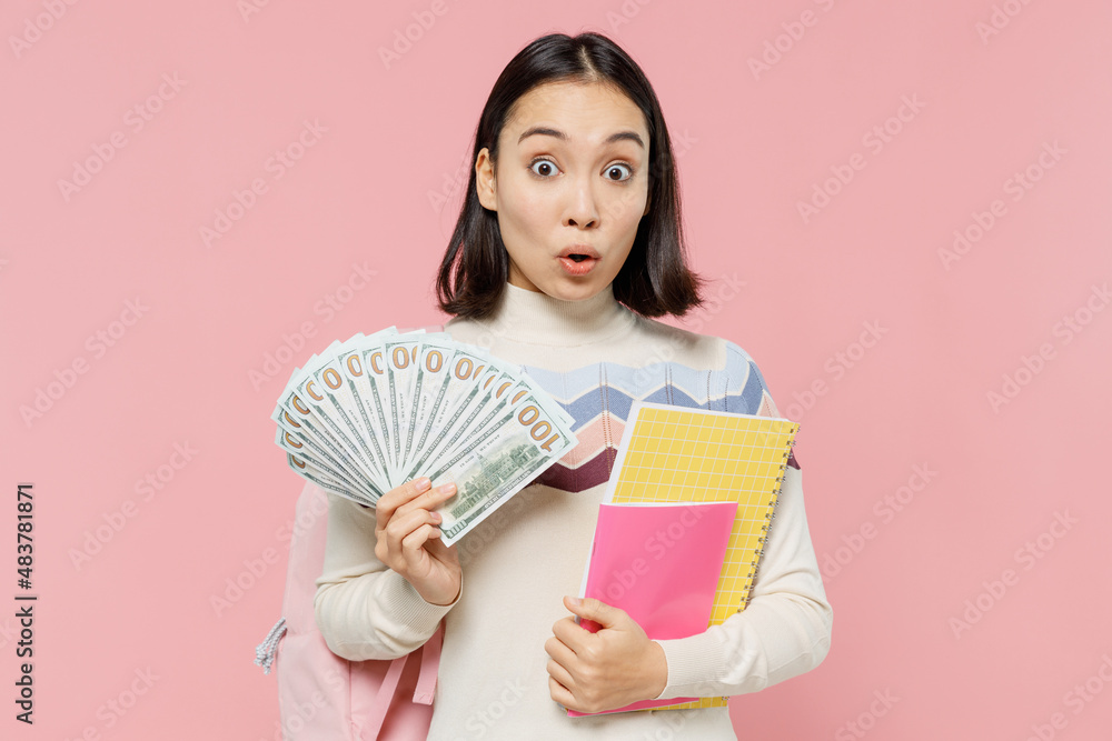Shocked teen student girl of Asian ethnicity in sweater backpack hold book fan of cash money in dollar banknotes isolated on pastel plain light pink background Education in university college concept Shocked teen student girl of Asian ethnicity in sweater backpack hold book fan of cash money in dollar banknotes isolated on pastel plain light pink background Education in university college concept