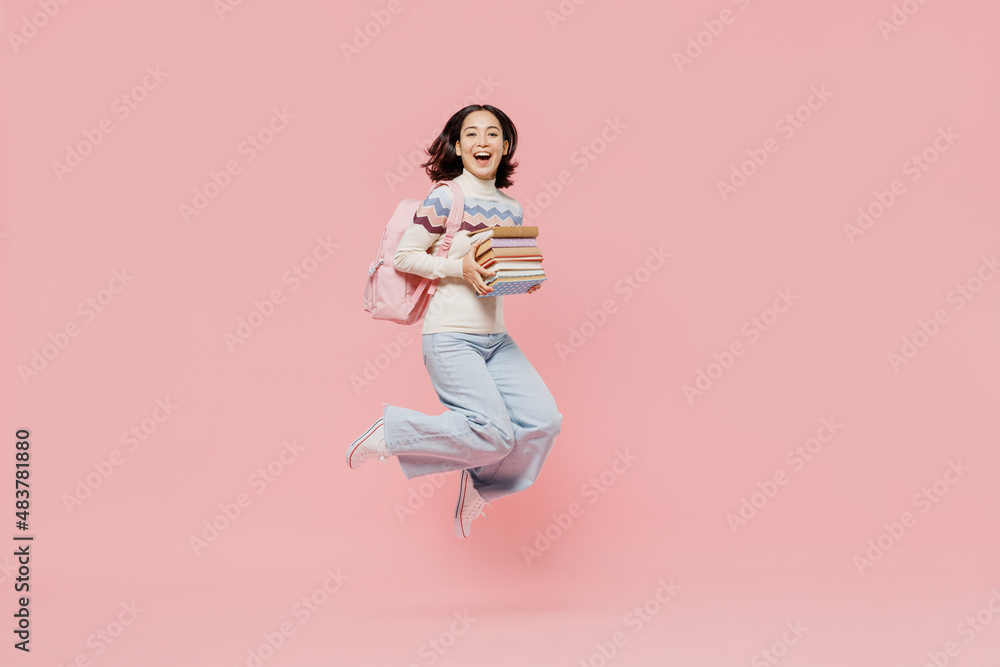 Full size happy excited teen student girl of Asian ethnicity wear sweater hold backpack jump high with pile of books isolated on pastel plain pink background Education in university college concept