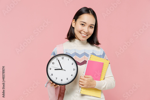 Smiling happy satisfied teen student girl of Asian ethnicity wear sweater backpack hold books clock isolated on pastel plain light pink background Education in high school university college concept.