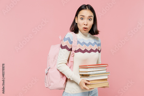 Shocked scared sad teen student girl of Asian ethnicity wearing sweater backpack hold pile of books look camera isolated on pastel plain light pink background Education in university college concept.