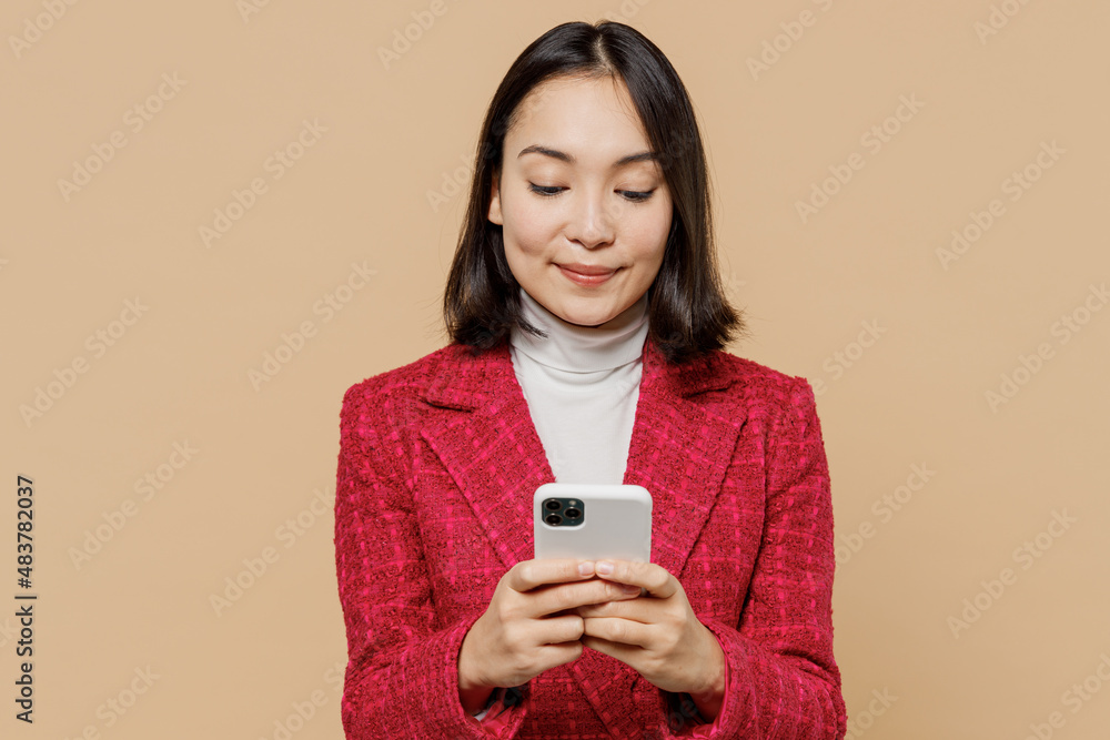 Smiling happy student woman of Asian ethnicity 20s wearing red jacket hold in hand use mobile cell phone isolated on plain pastel beige background studio portrait. People lifestyle fashion concept Smiling happy student woman of Asian ethnicity 20s wearing red jacket hold in hand use mobile cell phone isolated on plain pastel beige background studio portrait. People lifestyle fashion concept
