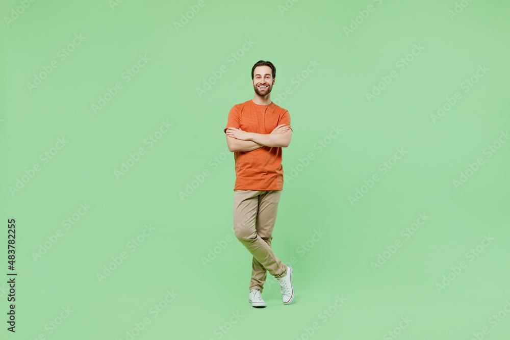 Full body young smiling fun man 20s wear casual orange t-shirt isolated on plain hold hands crossed folded look camera pastel light green color background studio portrait. People lifestyle concept. Full body young smiling fun man 20s wear casual orange t-shirt isolated on plain hold hands crossed folded look camera pastel light green color background studio portrait. People lifestyle concept.