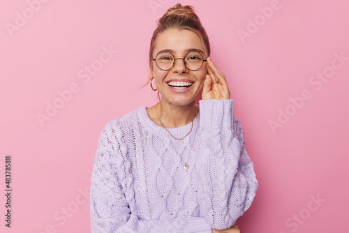 Portrait of pleasant looking woman smiles broadly shows perfect white teeth keeps hand on rim of spectacles dressed in knitted sweater isolated over pink background. People and positive emotions photo
