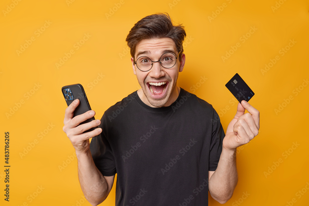 Positive handsome European man holds mobile phone and credit card makes online shopping or money transaction recommends banking dressed in casual black t shirt isolated over yellow background Positive handsome European man holds mobile phone and credit card makes online shopping or money transaction recommends banking dressed in casual black t shirt isolated over yellow background