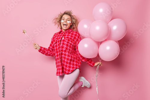 Joyful optimistic young woman dances with bunch of helium balloons comes to congratulate friend with anniversary exclaims gladfully holds magic wand wears beauty patches under eyes before party