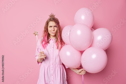 Frustrated unhappy long haired young woman holds bunch of inflated balloons and magic wand being angry with someone wears dress and crown poses against pink background doesnt like something.