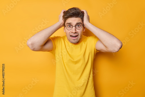 Shocked worried adult dark haired European man grabs head thinks about deadline looks stressed wears casual t shirt isolated over yellow background. People human reactions and emotions concept photo