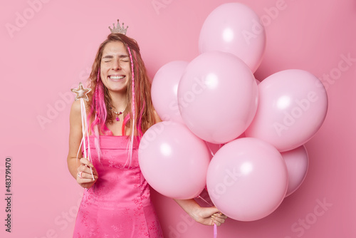 Joyful long haired young woman believes in miracle holds magic wand and bunch of inflated balloons makes wish on birthday wears festive dress isolated over pink background. Festivity concept