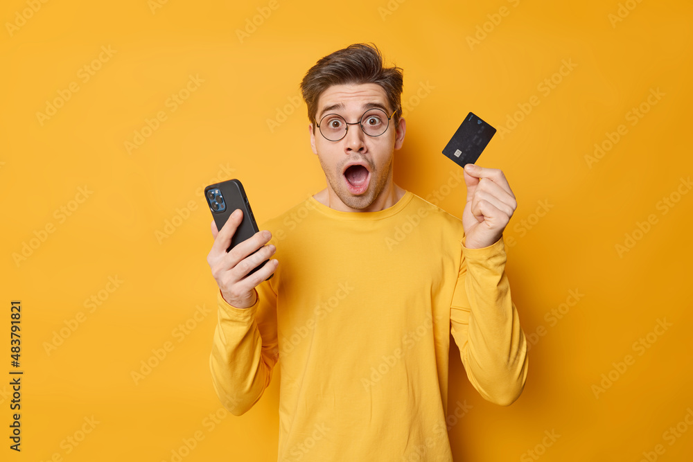 Shocked young man has financial probem with banking card uses mobile phone trie to solve shopping trouble wears spectacles and jumper isolated over yellow background makes money transaction. Shocked young man has financial probem with banking card uses mobile phone trie to solve shopping trouble wears spectacles and jumper isolated over yellow background makes money transaction.