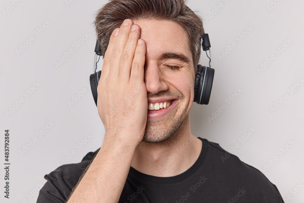 Headshot of happy brunet man covers half of face with palm feels joyful listens music via stereo headphones has good mood dressed in casual t shirt isolated over white background. Hobby concept Headshot of happy brunet man covers half of face with palm feels joyful listens music via stereo headphones has good mood dressed in casual t shirt isolated over white background. Hobby concept