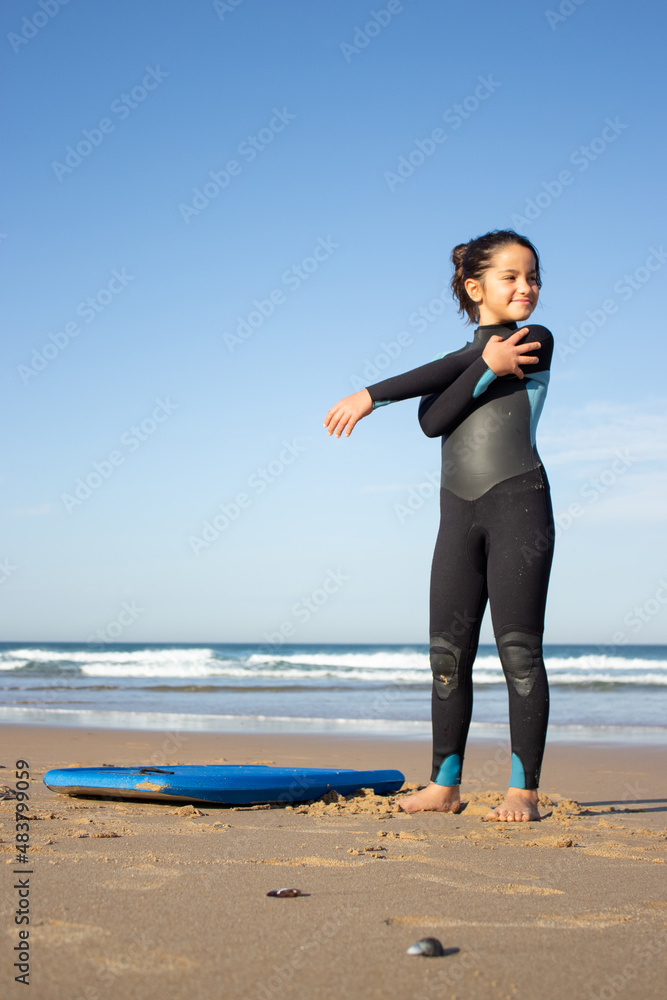 Smiling little girl exercising on beach. Dark-haired child in surf uniform on cloudy day, doing exercises. Sport, leisure, active lifestyle concept Smiling little girl exercising on beach. Dark-haired child in surf uniform on cloudy day, doing exercises. Sport, leisure, active lifestyle concept