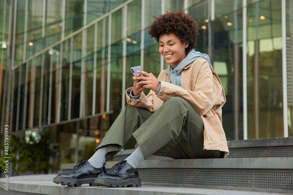 © wayhome.studio - Outdoor shot of happy curly haired millennial girl wears fashionable clothes sits on stairs near modern shopping centre reads message on smartphone enjoys recreation time. Modern technologies